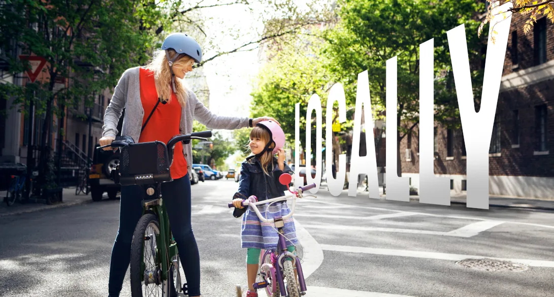 A parent and child with bicycles waiting at a city crosswalk, with the Locally wordmark set into the scene.